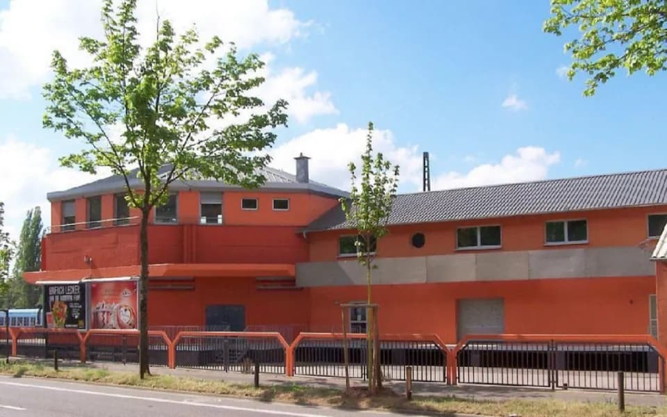 Straßenansicht der Gewerbe- und Gastronomieimmobilie in markantem Terrakotta-Orange mit polygonalem Kopfbau, Blechdach, Hallentrakt und eingefriedetem Vorplatz. Screenshot 2026-03-14 065023.jpg